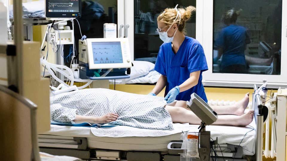 Eine Krankenschwester in blauer Krankenhaus-Kleidung steht mit Mundschutz am Bett einer Corona-Patientin auf der Intensivstation