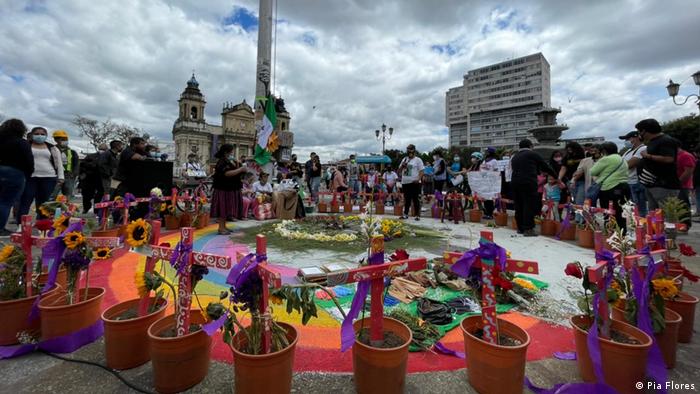 Auf einem Platz in Guatemala-Stadt stehen rund um einen mit Regenbogenfarben gestalteten Kreis kleine roten Blumentöpfen, in denen Sonnenblumen wachsen und in die rote Kreuze gesteckt sind. Proteste gegen Gewalt gegen Frauen in Guatemala.